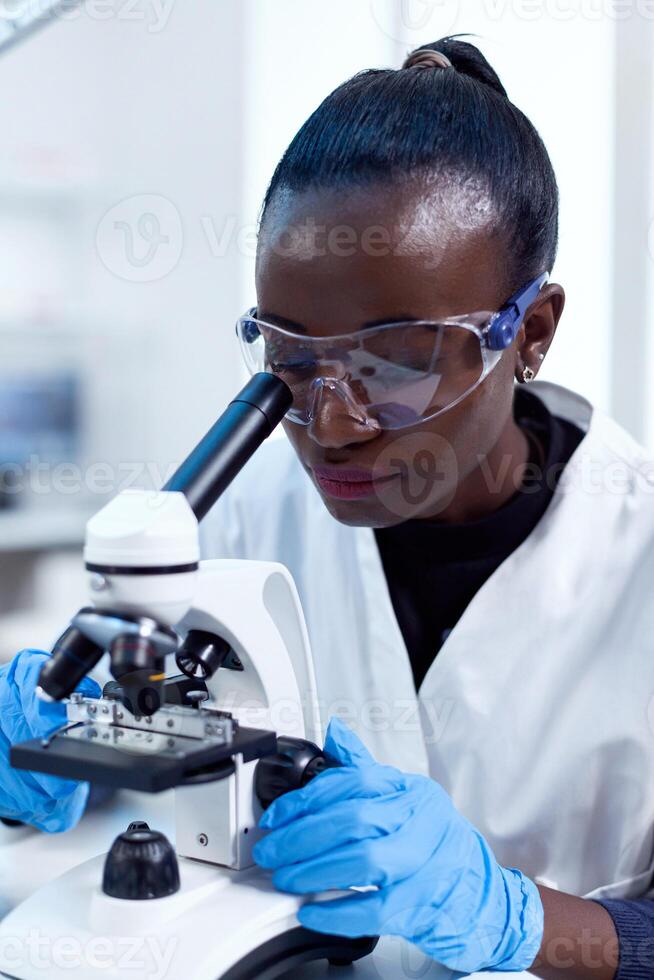 Close up of chemist of african ethnicity doing sample analysis looking through microscope. Black healthcare scientist in biochemistry laboratory wearing sterile equipment. photo