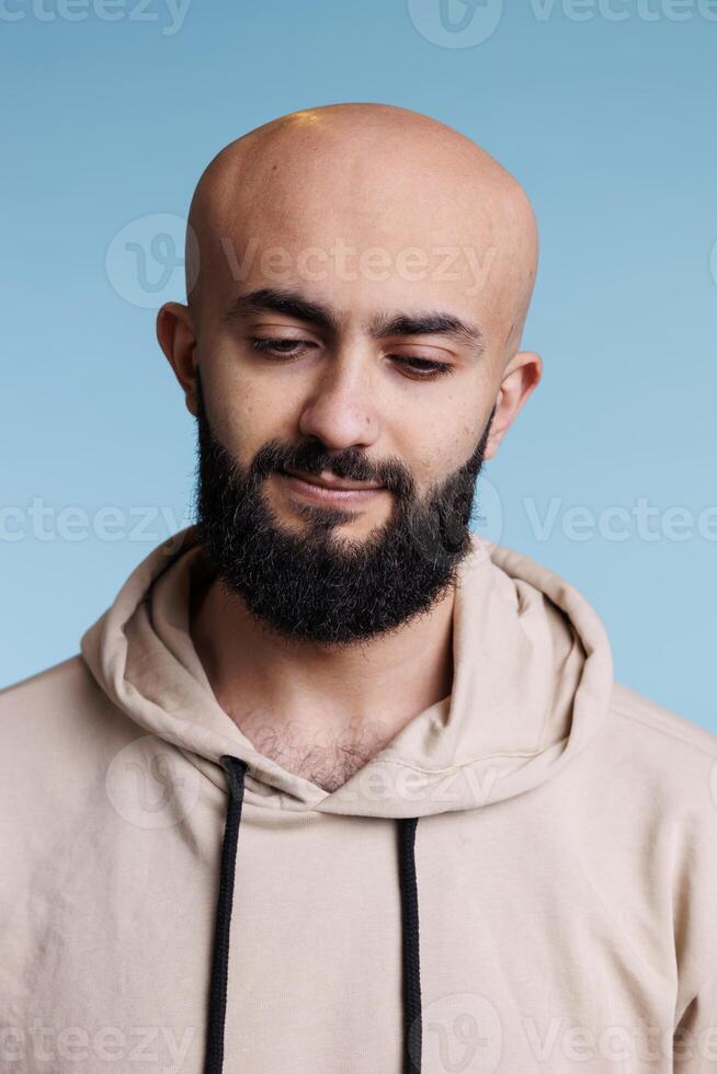 Young adult arab man looking down with thoughtful facial expression. Puzzled bald bearded person thinking, wearing casual beige hoodie and posing naturally on blue background photo