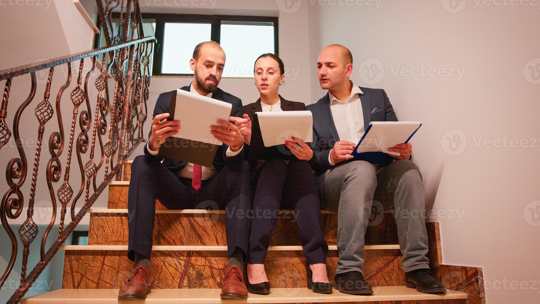 Project team of business workers looking on documents sitting on stairs explaining finance project doing overtime. Group of company manager and office executive on busy stairs of workplace. photo