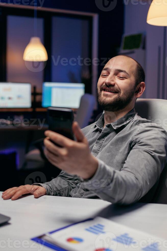 Cheerful entrepreneur saying hello talking on video call using phone. Businessman in the course of an important video conference while doing overtime at the office. photo