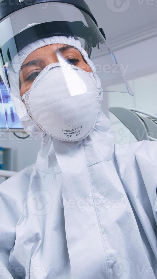 Patient pov of dentist holding dental tools wearing covid protection suit treating patient in new normal clinic. Stomatolog wearing safety gear against coronavirus during heatlhcare check of person. photo