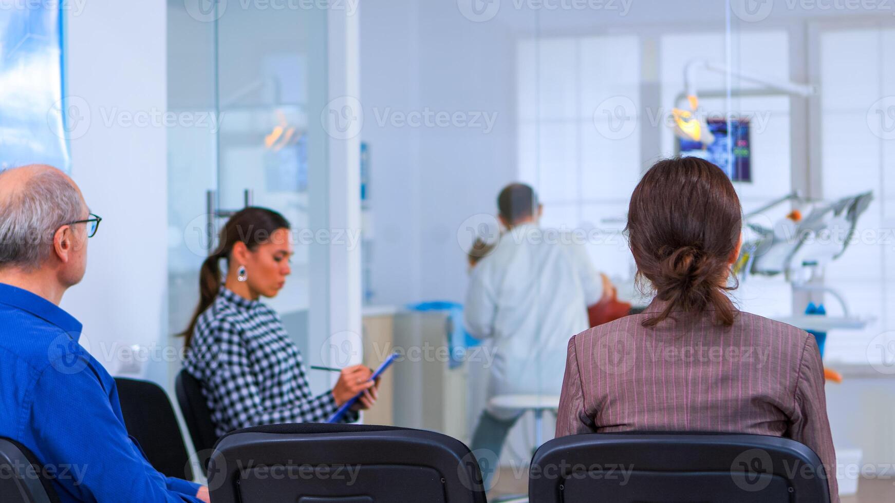 Back view of woman filling in dental form sitting on chiar in waiting room preparing for dental implants while doctor exemination patient in background. Crowded professional orthodontist office. photo