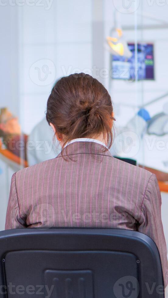 Back view of woman filling in dental form sitting on chiar in waiting room preparing for dental implants while doctor exemination patient in background. Crowded professional orthodontist office. photo