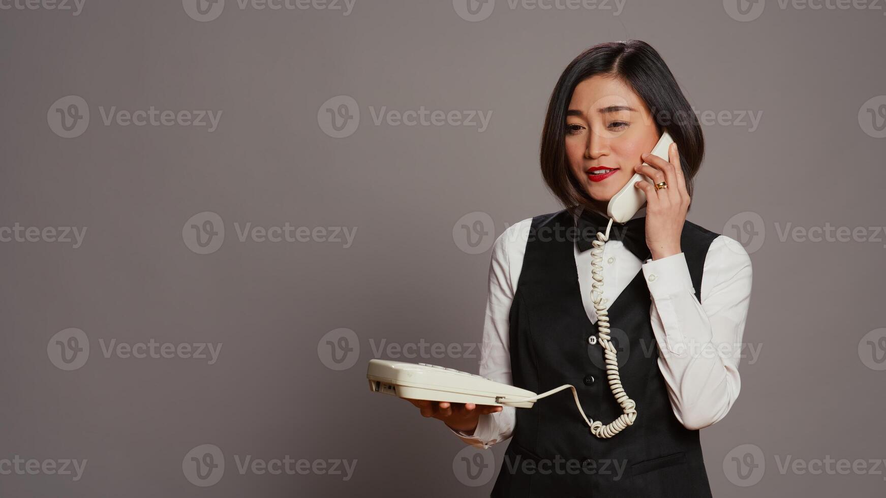 asiático recepcionista respondiendo teléfono fijo teléfono llamada en estudio, hablando en Clásico teléfono con cable. mujer frente escritorio personal utilizando teléfono con cable a confirmar habitación reserva. cámara b. foto