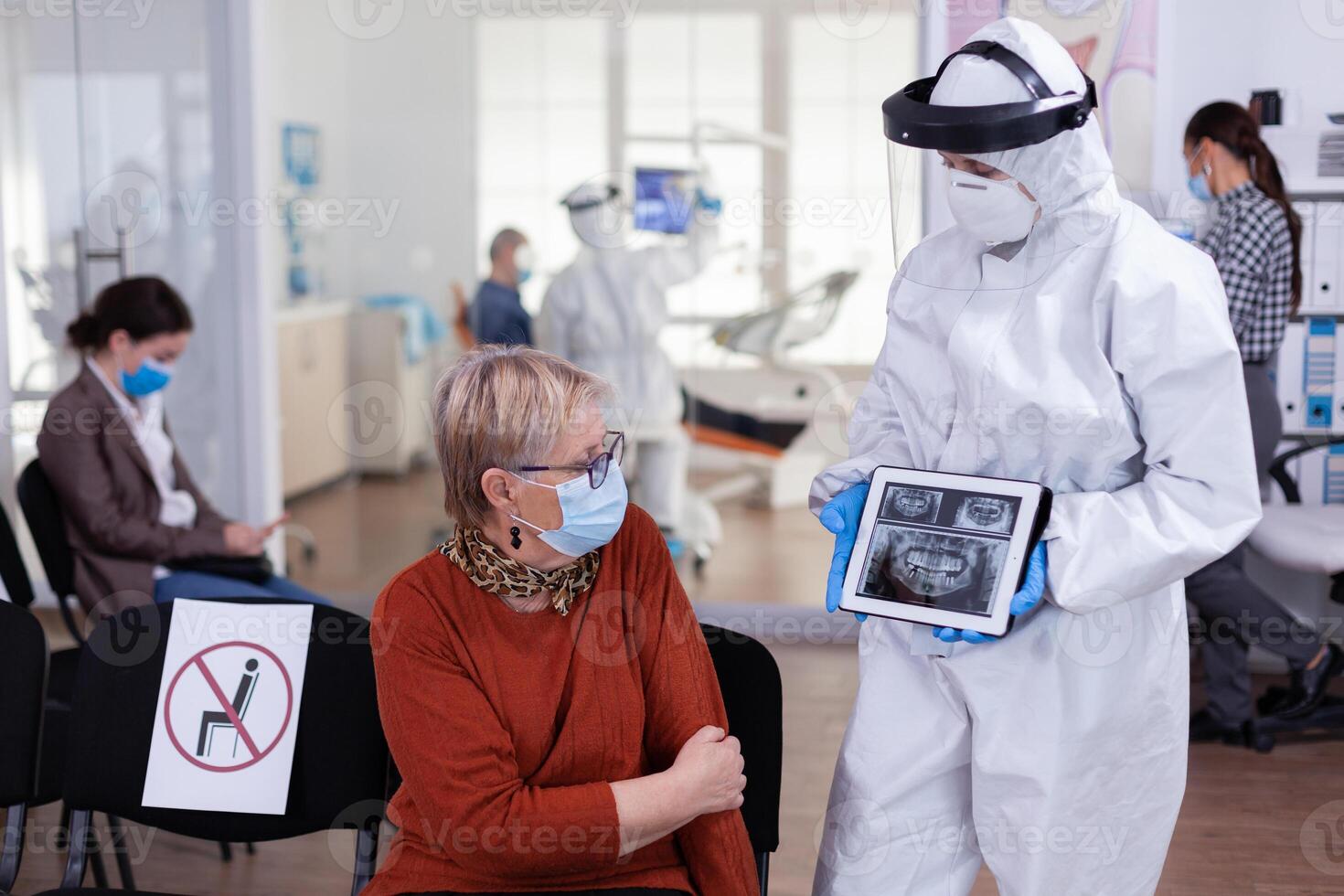 Dentist doctor with face shield pointing on tablet display explaining dental x-ray to senior patient during global pandemic. Nurse wearing protection suit, overall, mask and gloves, new normal. photo