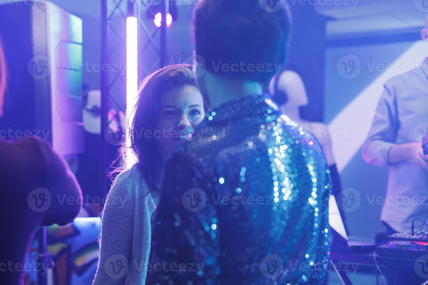 Woman partying and chatting with friend on crowded dancefloor in dark nightclub with spotlights. Cheerful girlfriends having fun together while attending discotheque in club photo