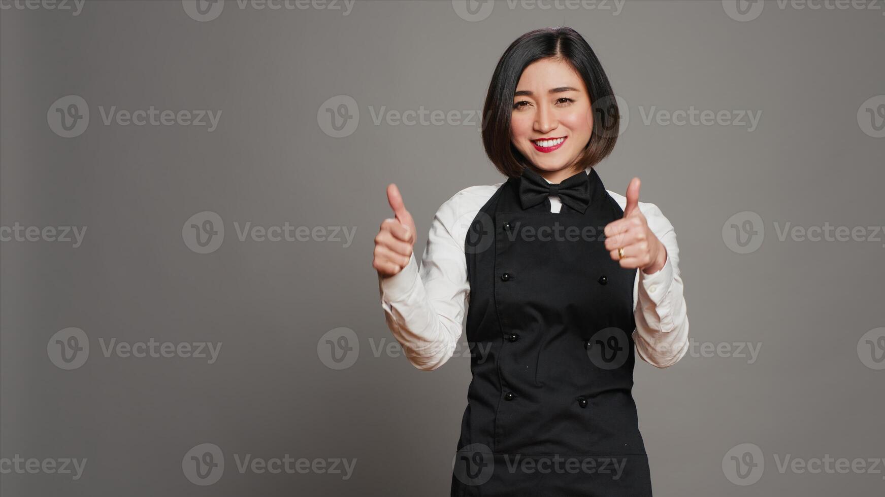 Smiling waitress giving thumbs up sign in front of camera, working in a five star restaurant to serve tables. Asian server with apron showing approval with okay symbol in studio, agreement. Camera A. photo
