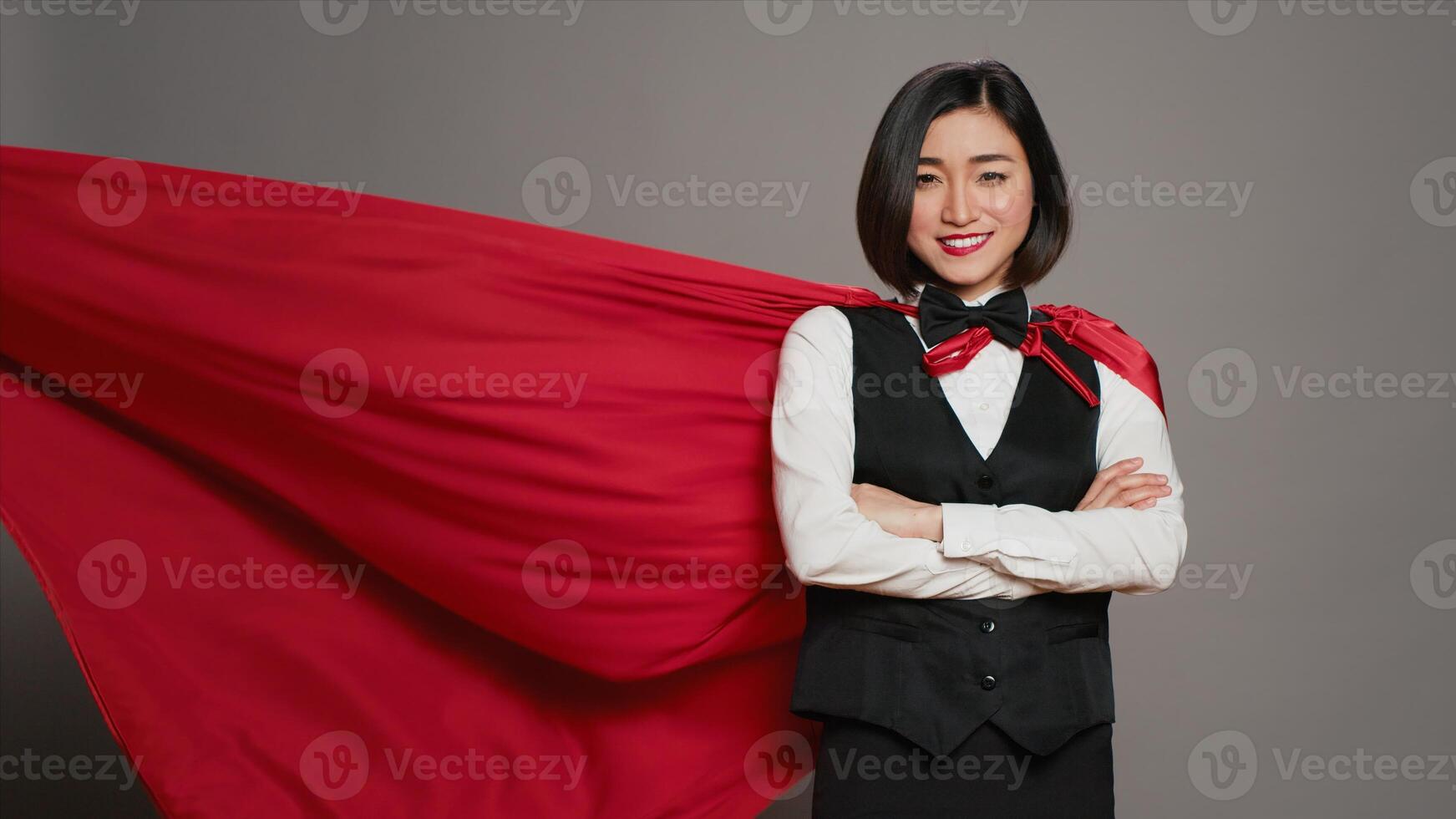 Asian receptionist posing with red superhero cape on camera, feeling confident and powerful while she wears a cloak as a hero. Hotel concierge operator standing with arms crossed. Camera A. photo