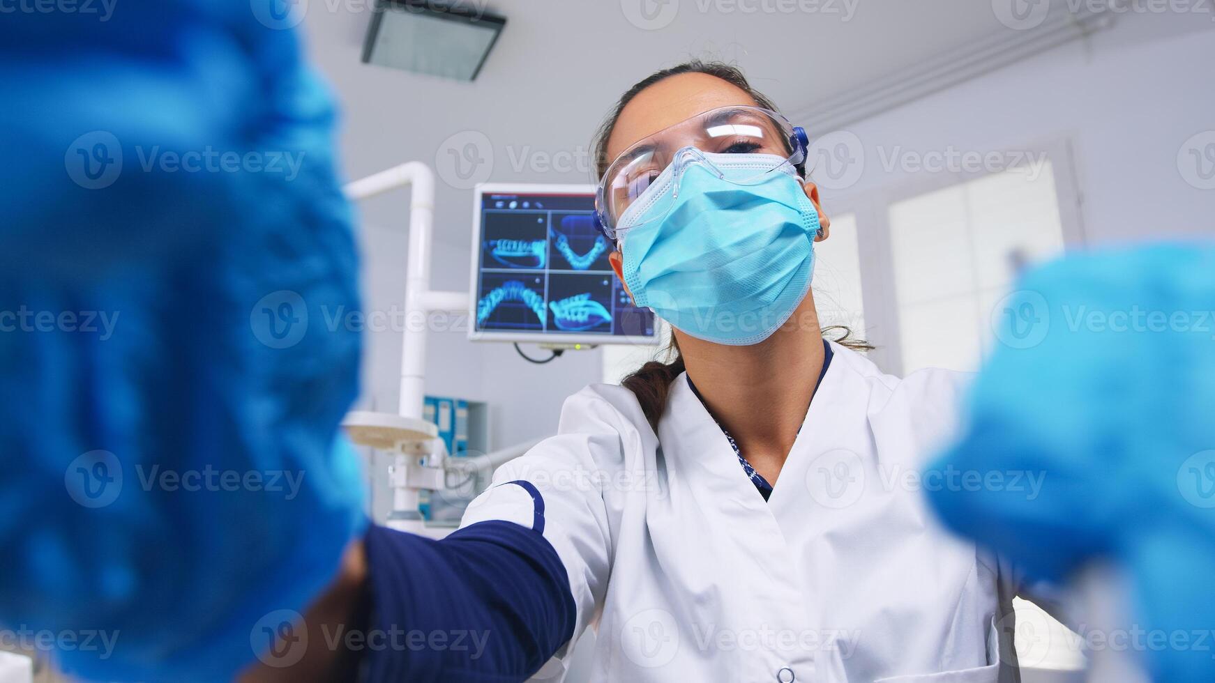 Patient point of view to dentist in protective mask holding tools examining person with toothache sitting on stomatological chair while nurse preparing tools for surgery. photo