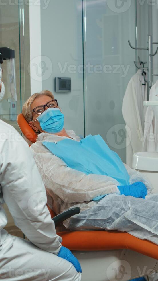Dentist doctor in coverall showing to senior patient x-ray on stomatological monitor during coronavirus. Concept of new normal dentist visit in coronavirus outbreak wearing protective suit and face shield photo