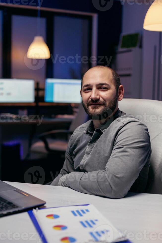 Focused freelancer smiling at camera resting hand on desk late at night. Smart businessman sitting at his workplace in the course of late night hours doing his job. photo
