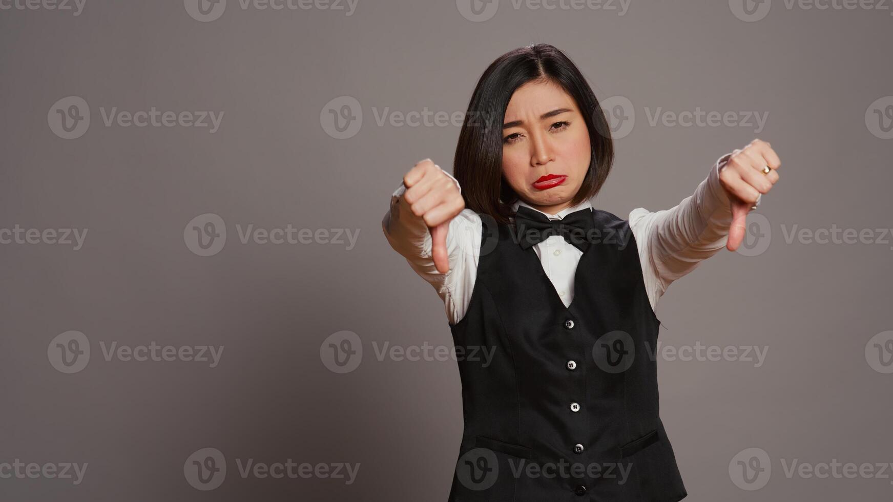 Receptionist showing thumbs down sign over grey background, expressing negativity and disapproval in studio. Woman hotel concierge giving dislike symbol, feeling displeased. Camera B. photo