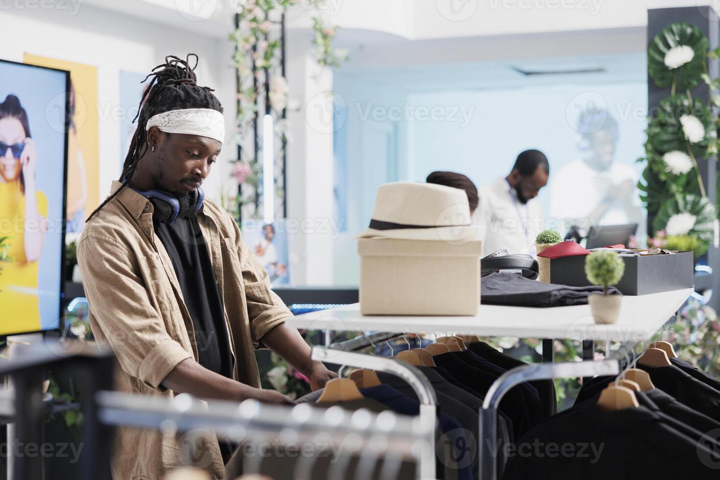 africano americano hombre hojeada estante con colgando ropa mientras compras en Moda boutique. joven comprador elegir elegante atuendo desde casual ropa de caballero nuevo colección en centro comercial foto