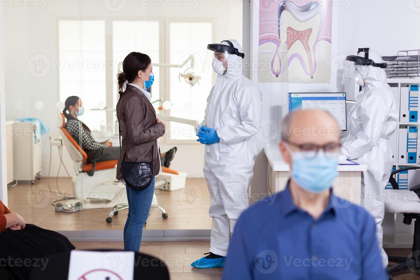 Dentist doctor dressed in ppe suit discussing with patient in stomatology reception corridor during appointment , in time of global pandemic with coronavirus health crisis. photo