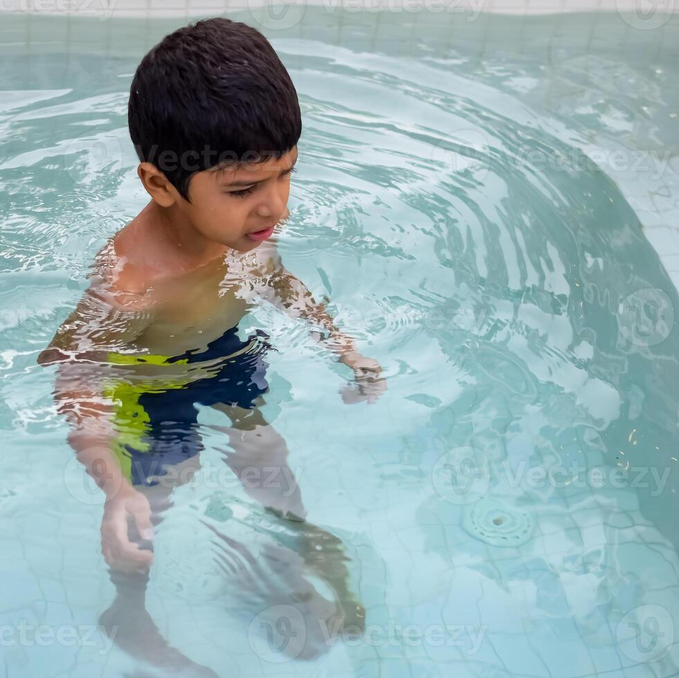 Happy Indian boy swimming in a pool, Kid wearing swimming costume along with air tube during hot summer vacations, Children boy in big swimming pool. photo