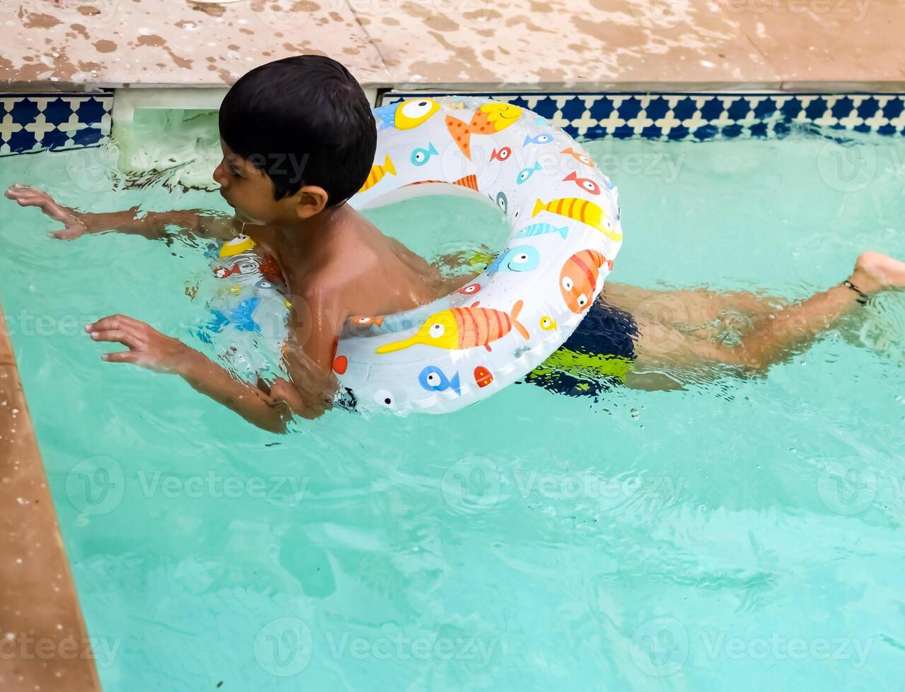 Happy Indian boy swimming in a pool, Kid wearing swimming costume along with air tube during hot summer vacations, Children boy in big swimming pool. photo