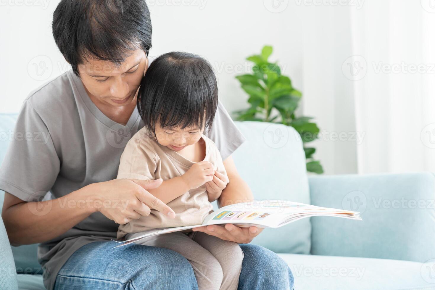 Happy Asian father relax and read book with baby time together at home. parent sit on sofa with daughter and reading a story. learn development, childcare, laughing, education, storytelling, practice. photo