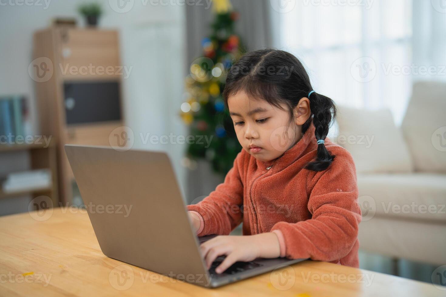 Asian Young Girl Focused on Using a Laptop at Home. A young girl with pigtails is engrossed in using a laptop on a wooden table, with a Christmas tree in the background. photo