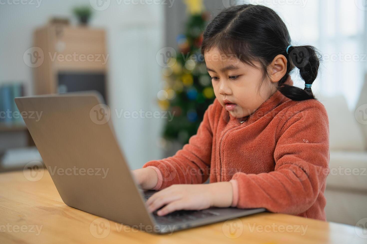 Asian Young Girl Focused on Using a Laptop at Home. A young girl with pigtails is engrossed in using a laptop on a wooden table, with a Christmas tree in the background. photo