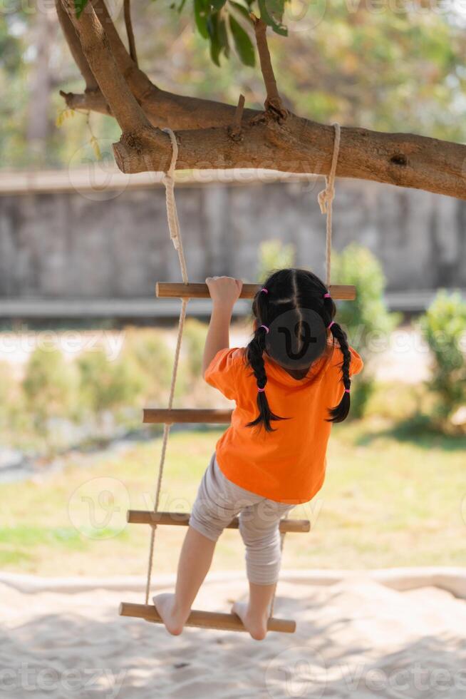 Back view. Young Girl Playing on a Rope Ladder. A little girl in an orange shirt climbs a rope ladder at a playground, with concentration. Baby girl kids activity concept. photo