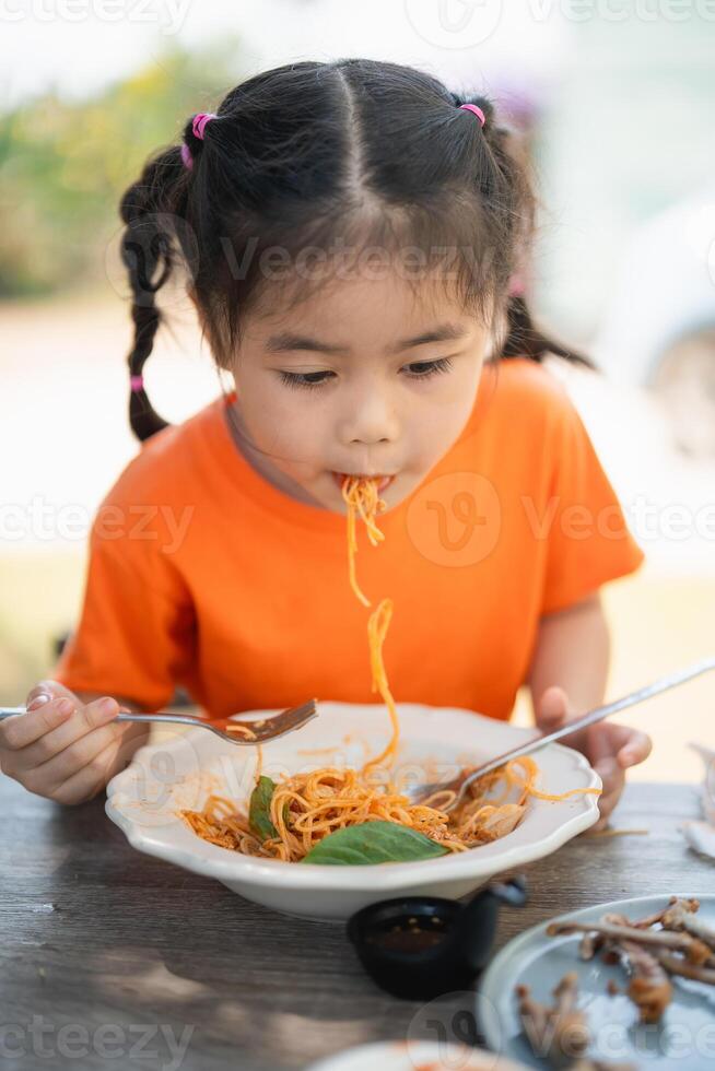 Young Girl children Enjoying a Forkful of Spaghetti. Close-up of a little girl in an orange shirt eating spaghetti with a focused expression, outdoors restaurant. photo