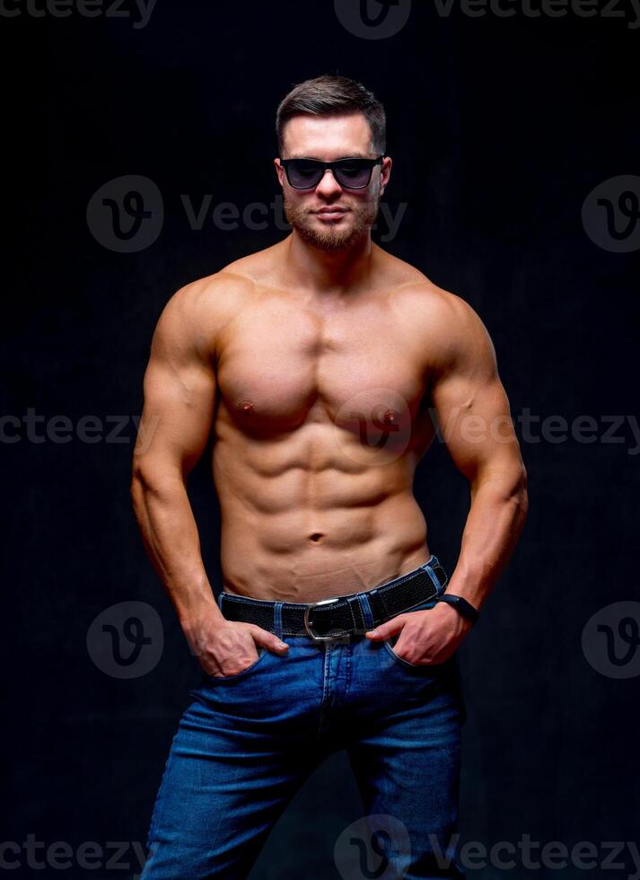 A portrait of a hot handsome strong guy man without a shirt against dark background. hands in pockets wearing glasses. Studio photo. Male beauty concept. Closeup. photo