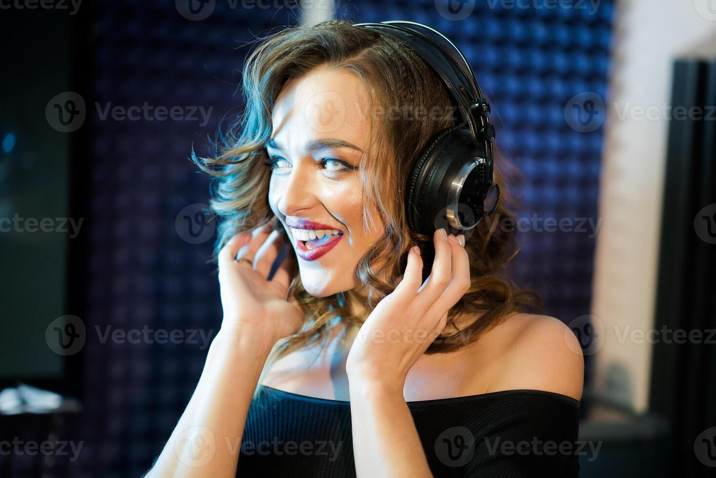 hermosa sonriente joven mujer con ondulado pelo conmovedor auriculares por su manos y mirando lado. contento hembra cantante en un profesional música estudio. de cerca foto