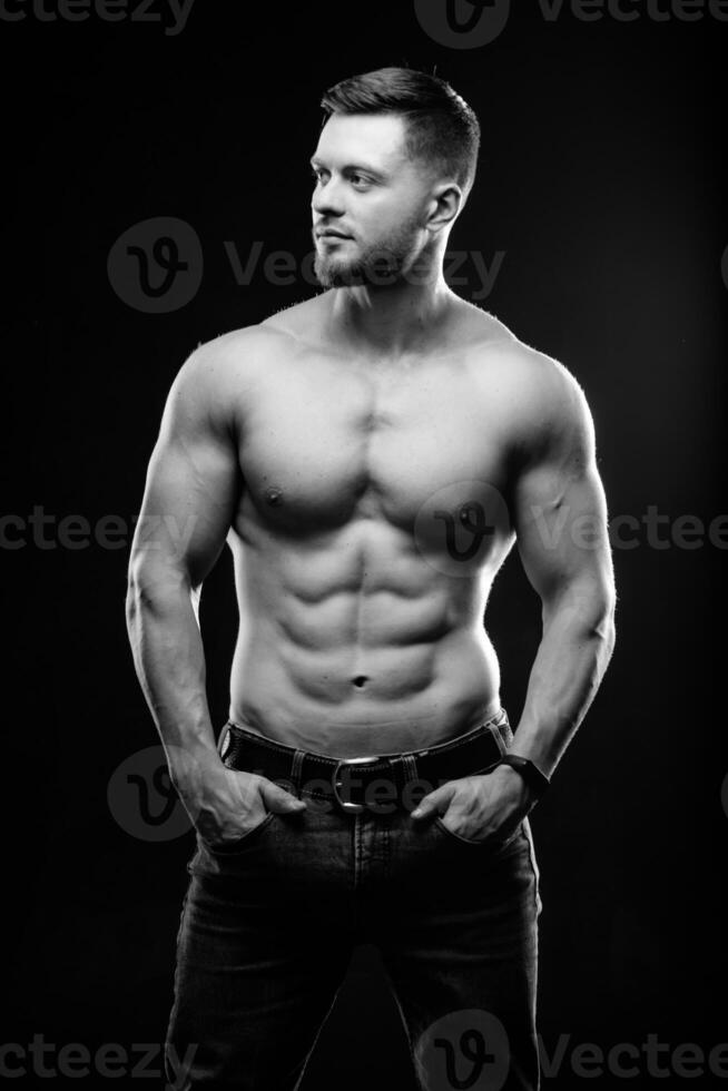 Muscular guy with naked torso posing with hands in pockets. Studio photo. Portrait of a handsome man in jeans on dark background. Black and white photo. Closeup. photo