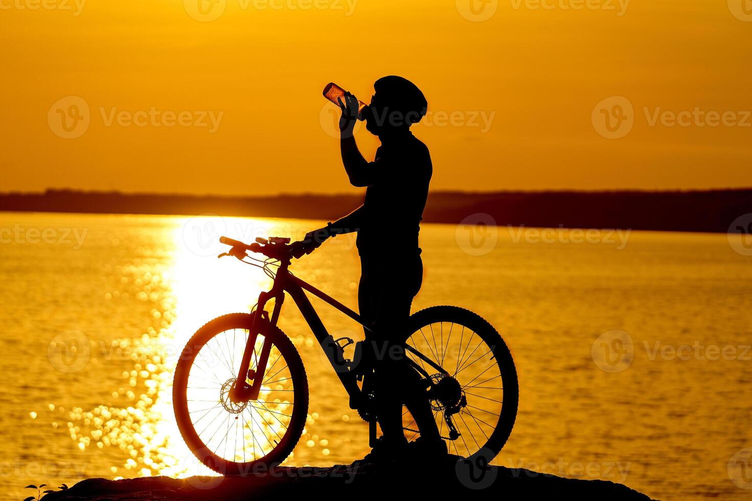 Silhouette of cyclist. Sporty guy cycling in helmet drinks water on the natural background of setting the sun in the evening photo