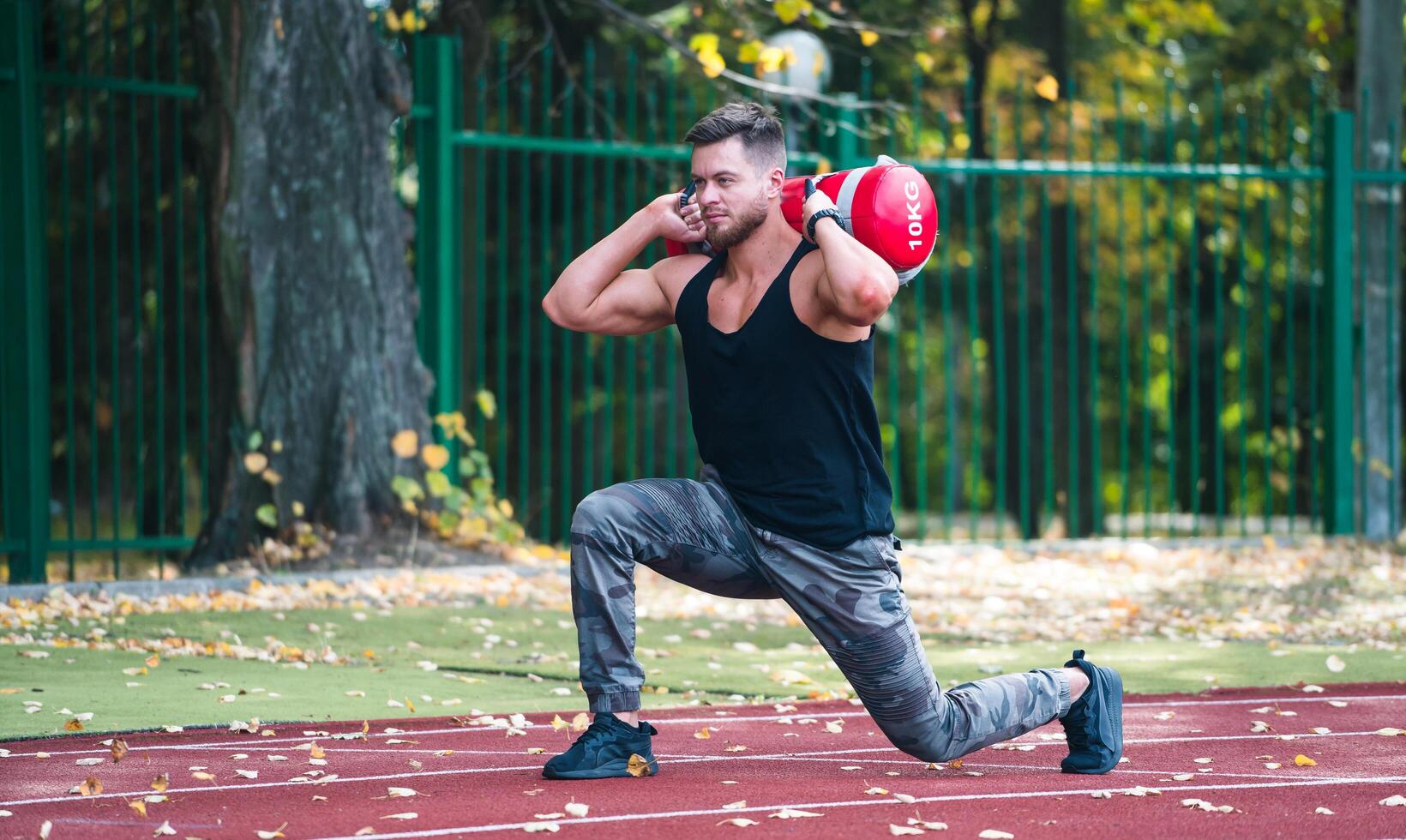 hermoso joven atleta trabajando fuera en un corriendo pista. joven hombre haciendo sentadillas con un pesado 10 kilo bolso en el pista. deporte y bienestar concepto. de cerca foto