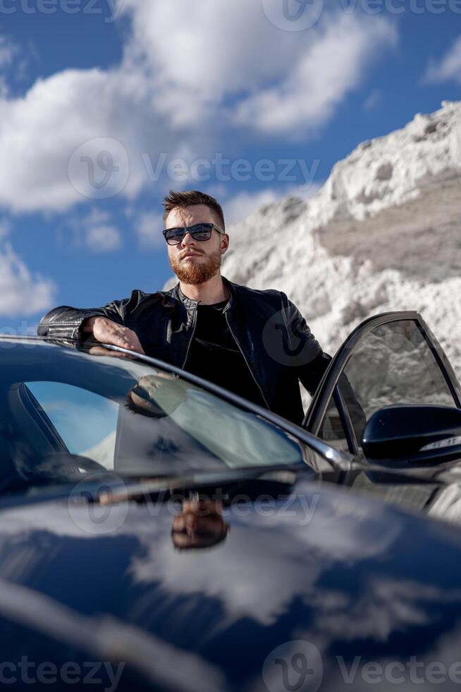 atractivo hombre en pie cerca abrió coche puerta. bonito ver de montaña y cielo. elegante combinación de negro y blanco. foto