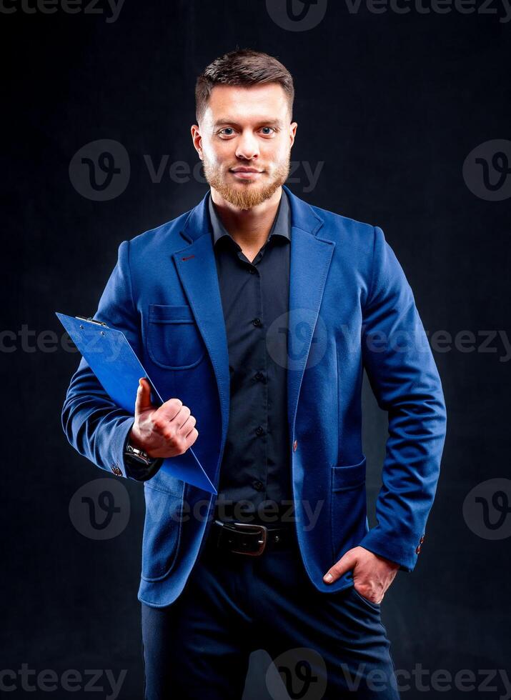 Half-length portrait of handsome young smiling man wearing dark shirt and blue suit holding blue folder, looking at camera. Black background. Money and business concept. photo