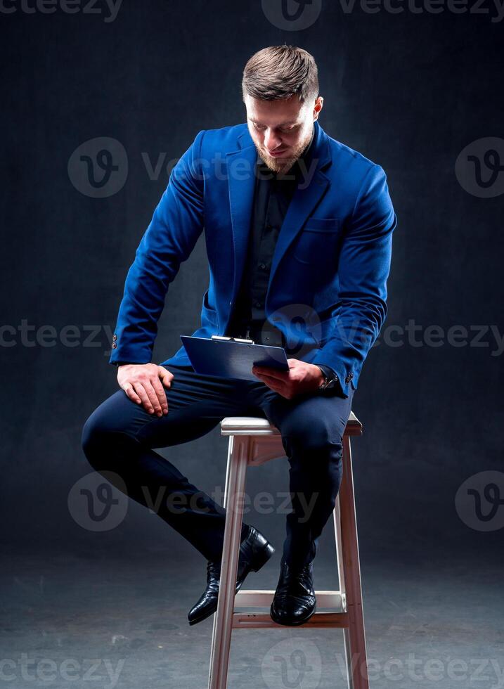 Young man with folder, isolated on dark background sitting on a chair reading notes. man wearing suit. photo