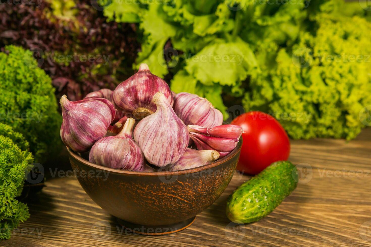 A full bowl with garlic bulbs. Tomatoes and cucumbers on a wooden table. Green salad in the background. photo