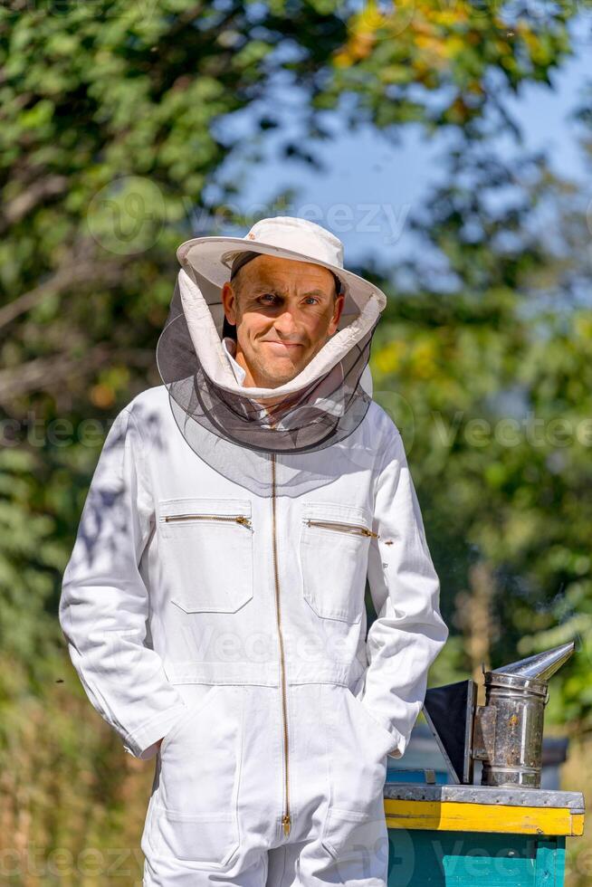 Adult man in protective uniform standing near beehives. Beekeeper doing his work with honey crop. photo