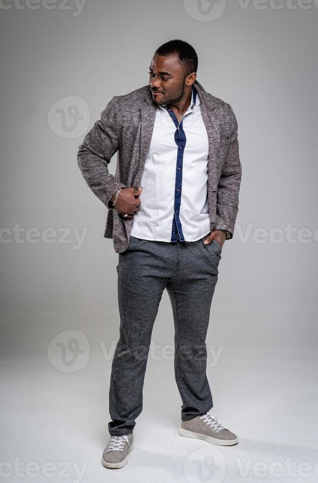 Smiling african american man posing with one arm in pocket and holding stylish jacket. Attractive business man in fashionable grey suit and black tie looking away. Full-lenght photo. photo