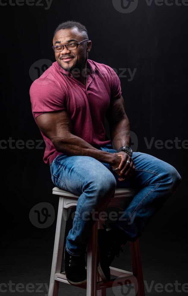 joven hermoso hombre en rojo camisa y pantalones sentado en silla. atlético muscular masculino posando para cámara en silla. foto