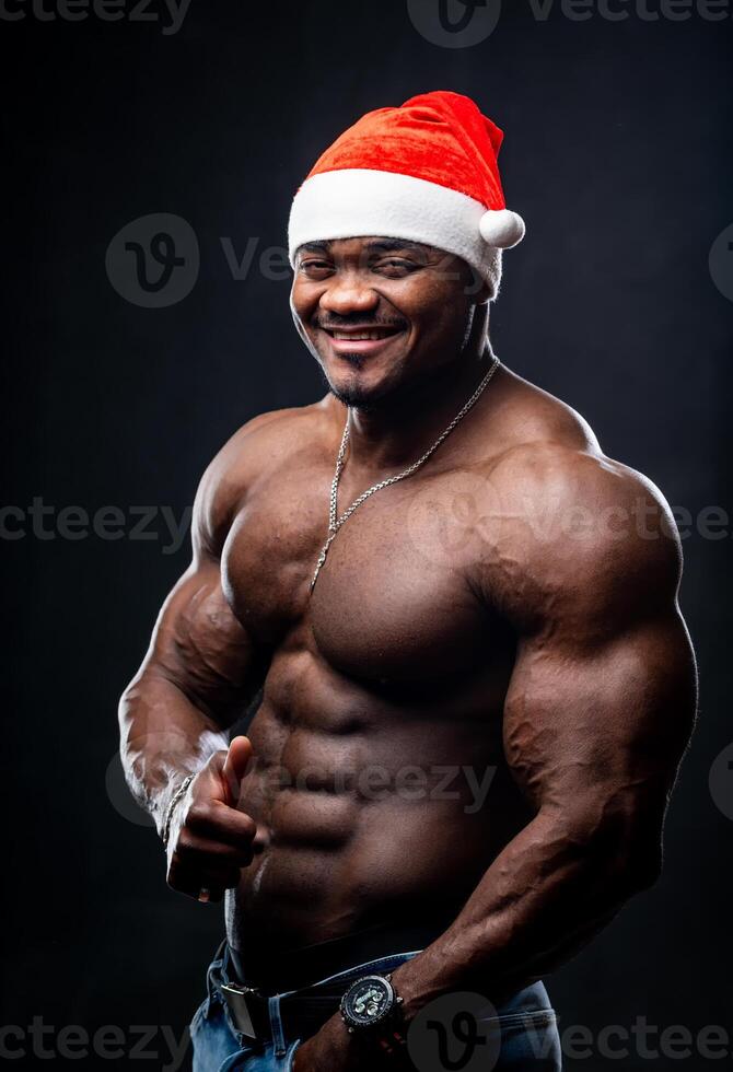 Happy sexy Santa Claus in red hat posing on black background. Muscular bodybuilder with naked torso looking at camera. photo