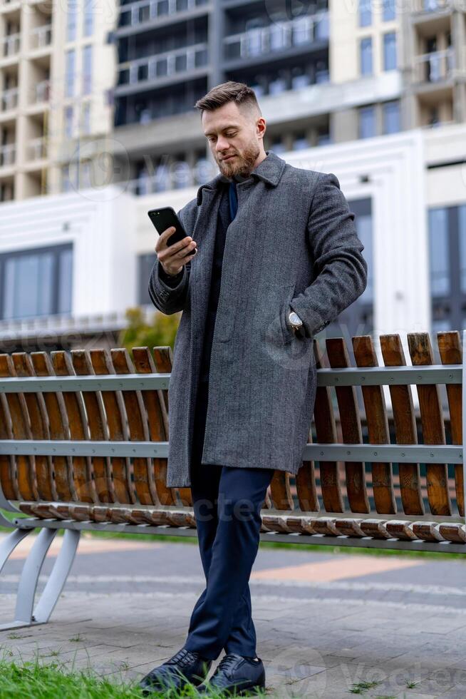 Adult man in suit standing near bench with phone. Businessman focused on phone work or job. photo