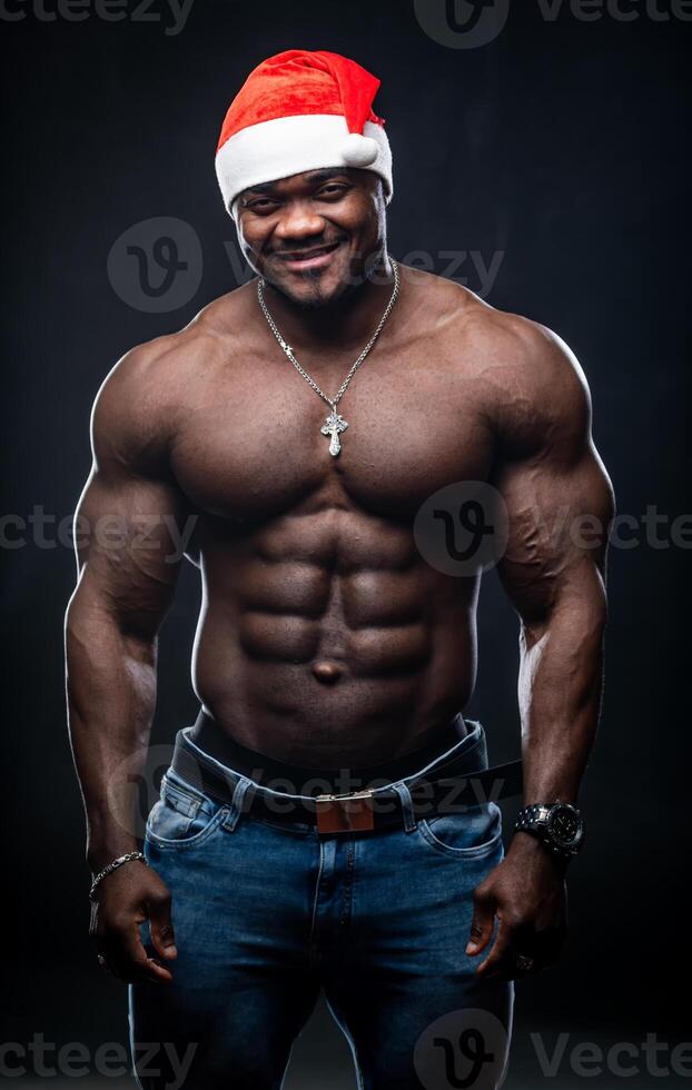 Happy muscular bodybuilder in Christmas hat. Funny handsome naked Santa Claus posing on black background looking at camera. photo