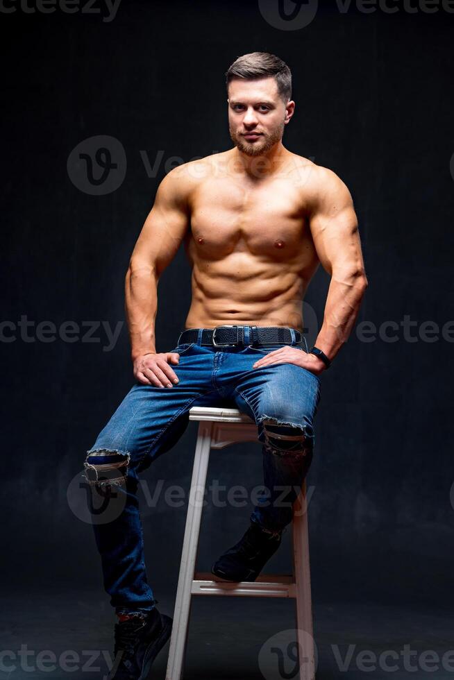 Muscular and fit young bodybuilder fitness male model posing on chair. Studio photo. Full size portrait. photo