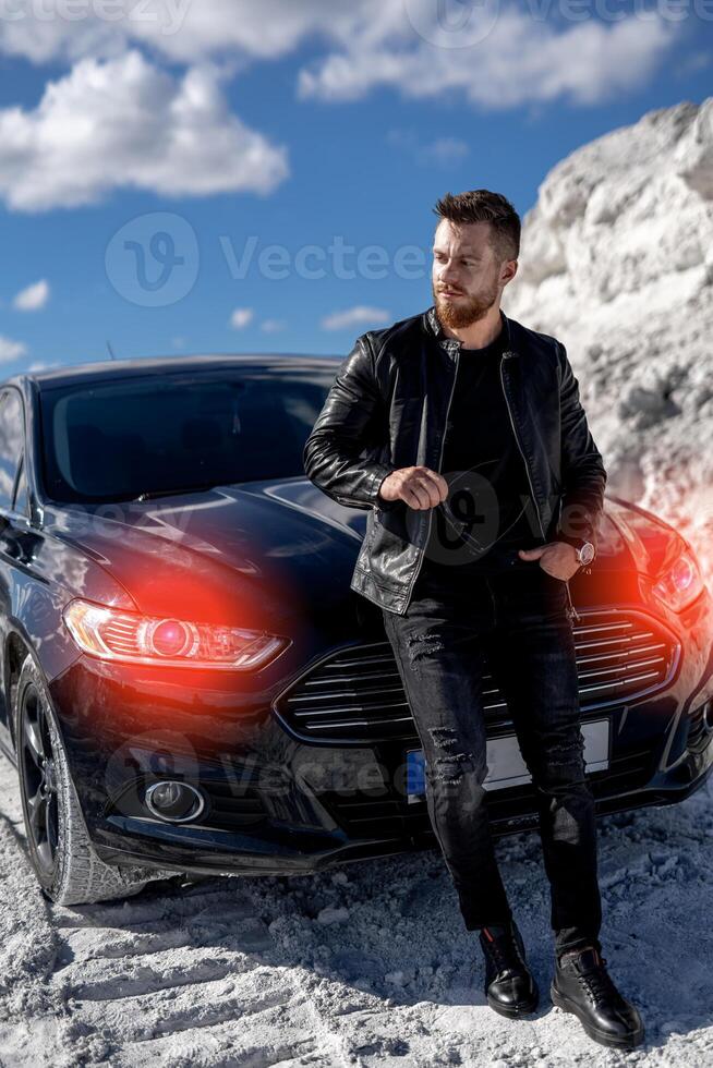 Man in black stylish clothes standing near black car. Man holding sunglasses. Beautiful location. photo