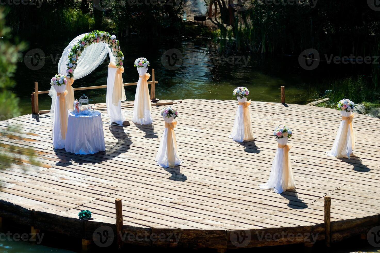 Wedding arch beautifully decorated and a small table, bouquets of flowers prepared for ceremony outdoors. Wooden surface in the form of a circle over the river for wedding event. photo