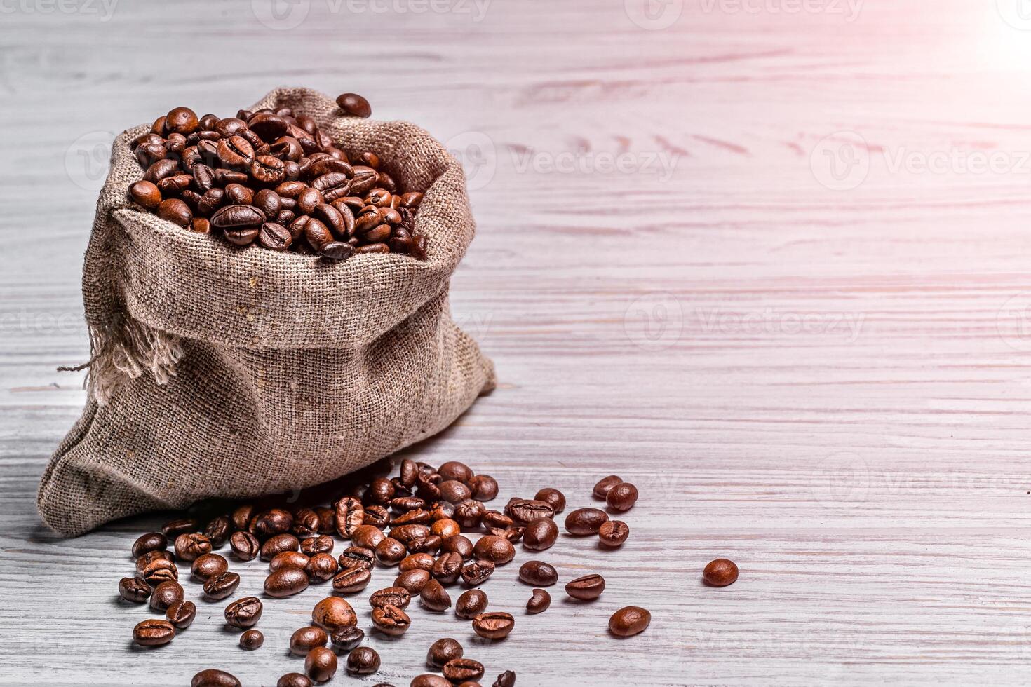 Small sack of coffee beans and some grains lying near it on the light background. Coffee beans scattered near the bag over the wooden table. photo