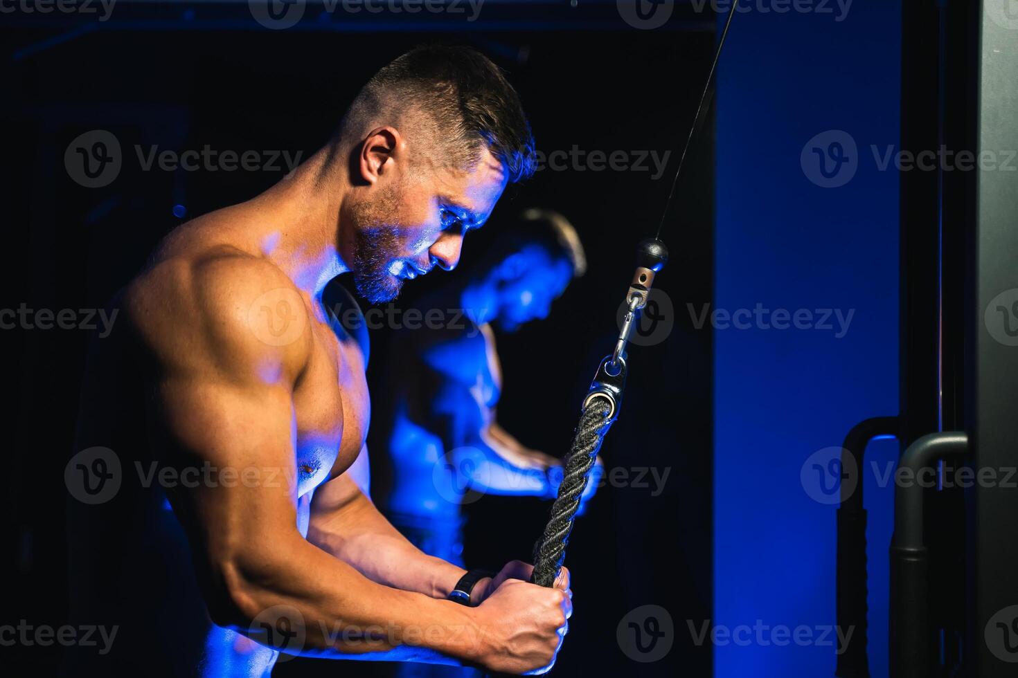 Handsome brutal sexy strong bodybuilder athletic fitness man pumping up muscles and doing workout with trx. Bodybuilding concept. Black and blue light background. View from the side closeup. photo