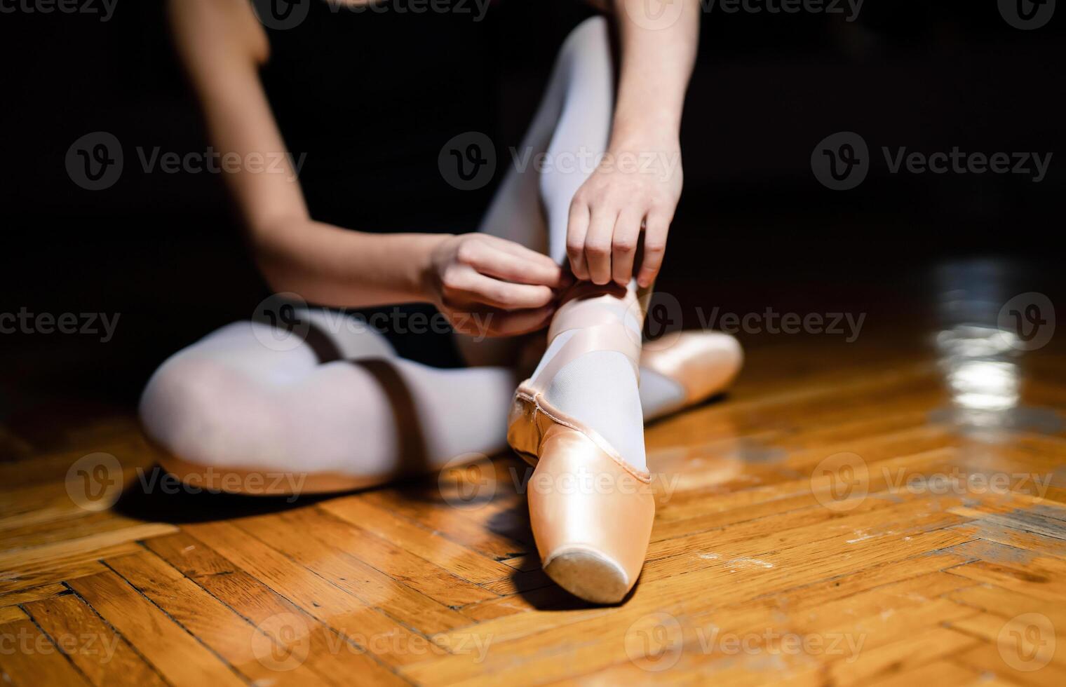 Unknown ballerina is tying the ribbon of pointe shoes on the wooden floor in a ballet class. The ballerina ties the pointes on slender legs. Close-up photo