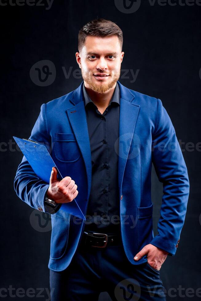 Handsome man in business casual clothing standing holding a clipboard, in front of a dark background. Portrait. photo