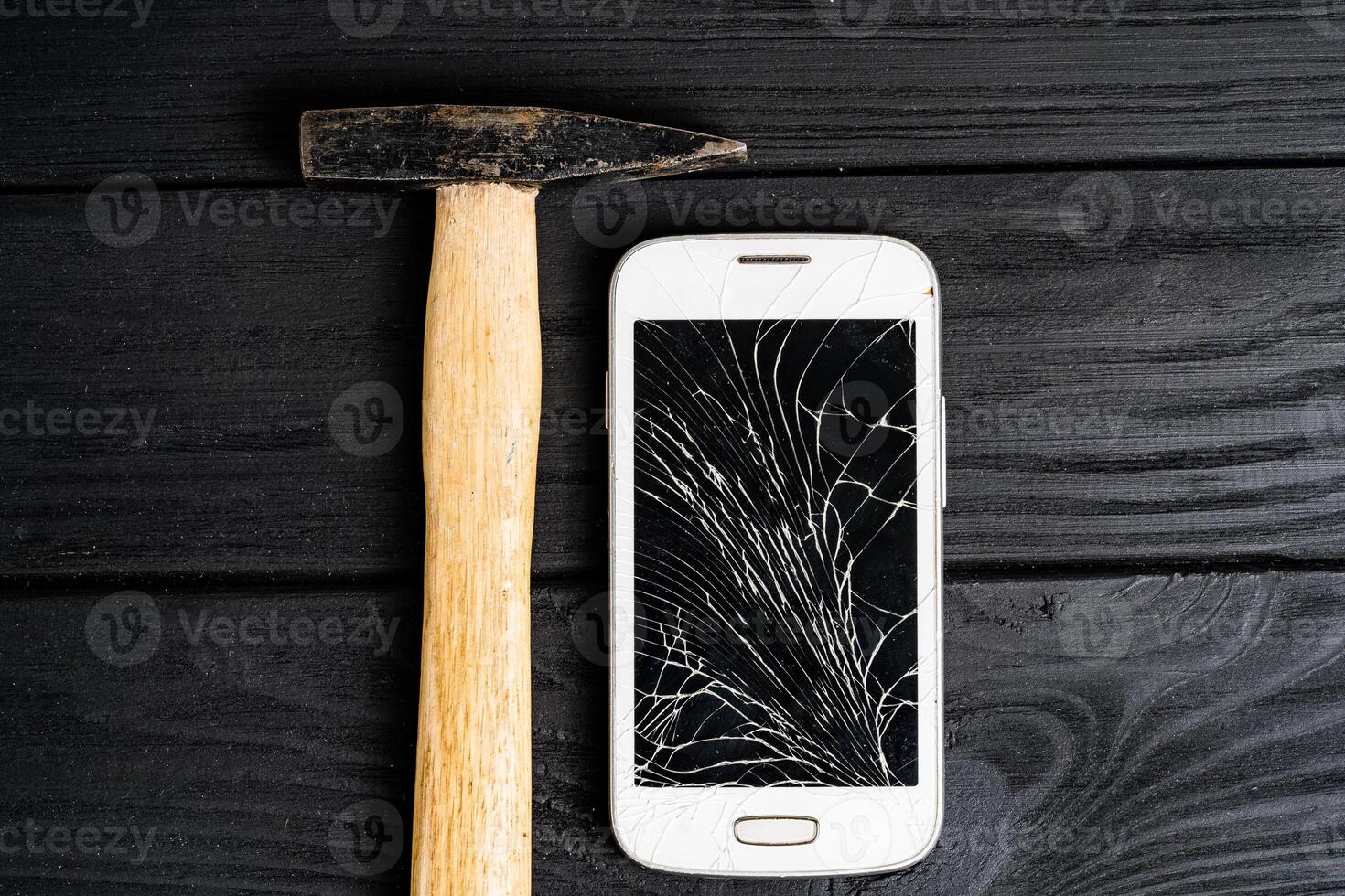 Smartphone smashed by hammer isolated on black wooden background. Broken touchscreen of a smartphone lying close to the hammer on table. photo