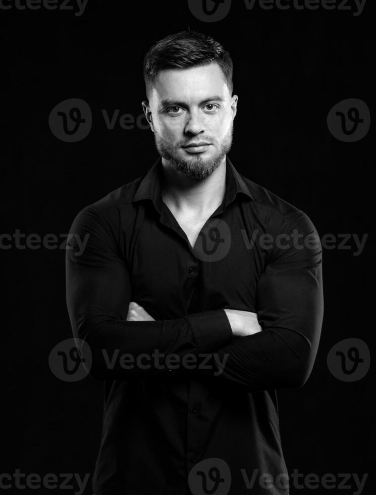 Handsome man in black shirt smiling isolated on dark background. Standing cross hands. Muscular male, athletic. Fashion portrait. Black and white photo