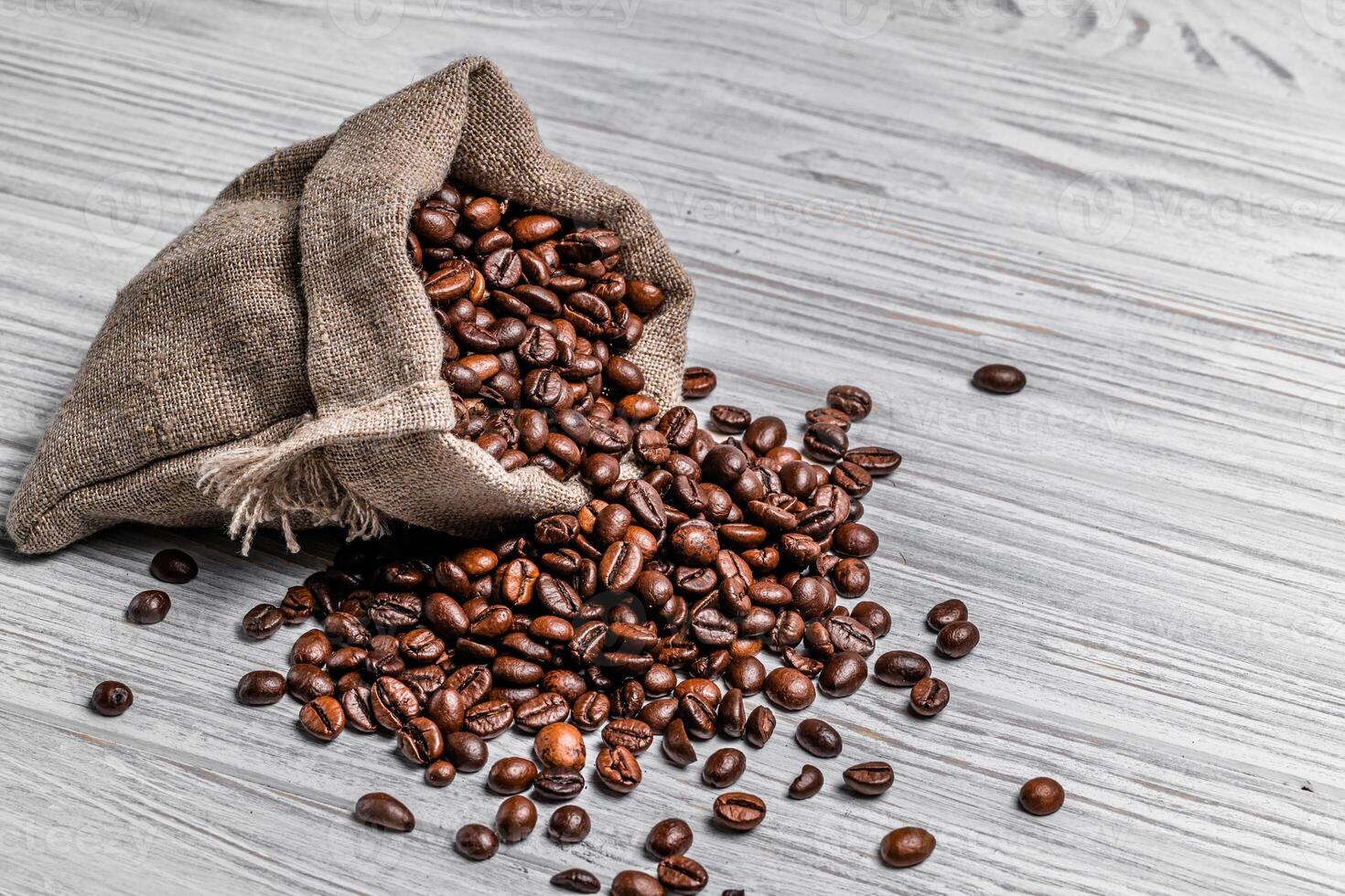 Bag of natural brown coffee beans and some scattered seeds on the light wooden background. Roasted coffee grains spilling out of sack on the table. photo